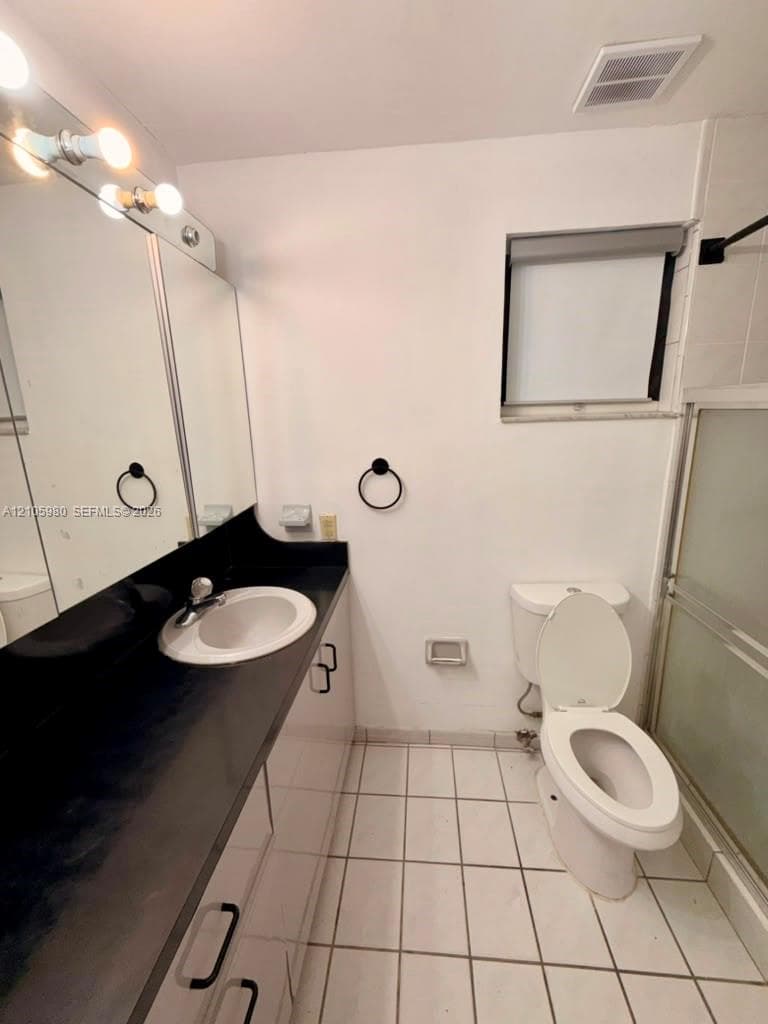 Bathroom featuring a vanity with dark solid-surface countertop, integrated basin, large wall-to-wall mirror, tiled flooring, and a glass-enclosed shower