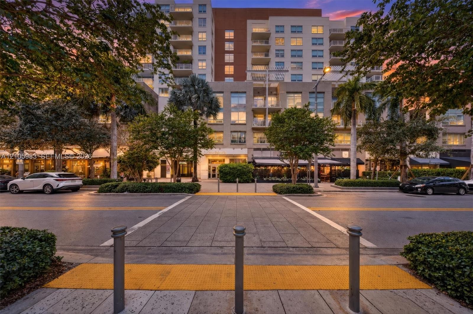 Multi-story building exterior with private balconies, ground-level retail frontages, decorative bollards, and mature landscaping