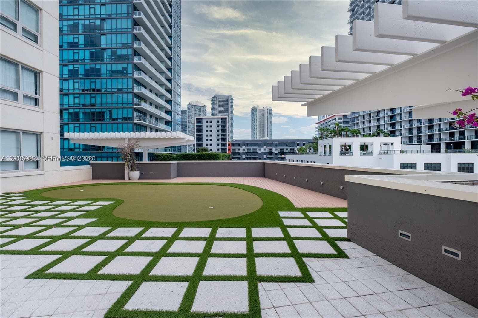 Outdoor terrace featuring a synthetic putting green, white paver grid, architectural pergola structures, and recessed wall lighting
