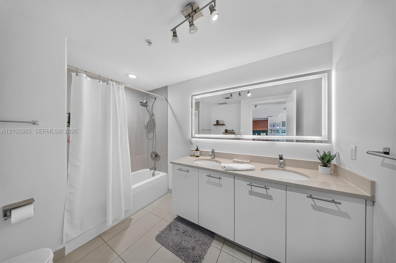 Double vanity featuring integrated sinks, light-toned solid surface countertops, flat-panel cabinetry, a large illuminated mirror, and a tub-shower combination with tile surround