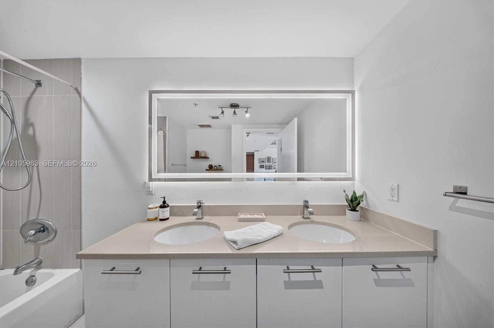 Dual-sink vanity with a solid-surface countertop, white cabinetry with horizontal hardware, backlit rectangular mirror, and a tile-surround shower and tub combination