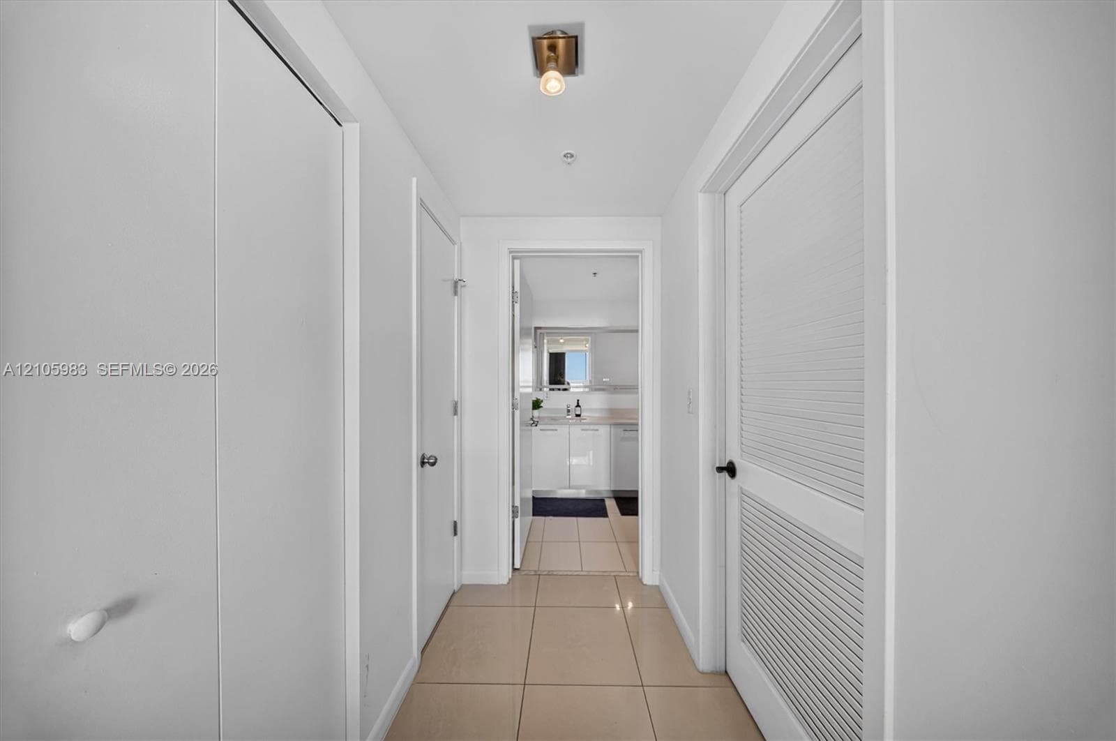 Interior hallway featuring large-format tile flooring, a brass-finished flush-mount light fixture, and louvered panel doors with matte black hardware
