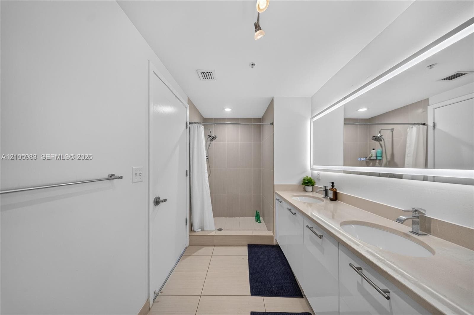 Bathroom featuring a dual-sink vanity with solid-surface countertops, large wall-to-wall backlit mirror, and a walk-in shower with tiled surround