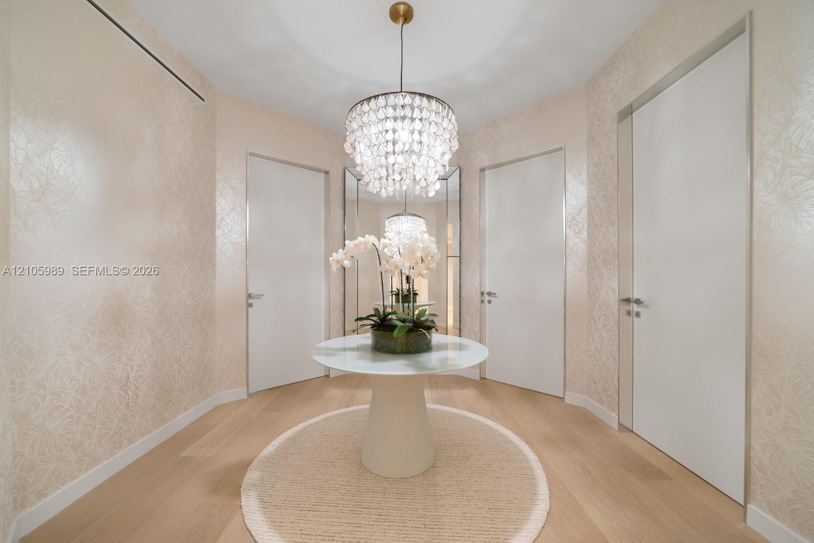 Foyer featuring light-toned wood-finish flooring, textured wall coverings, a tiered crystal chandelier, full-height flush doors with metallic casing, and floor-to-ceiling mirror panels