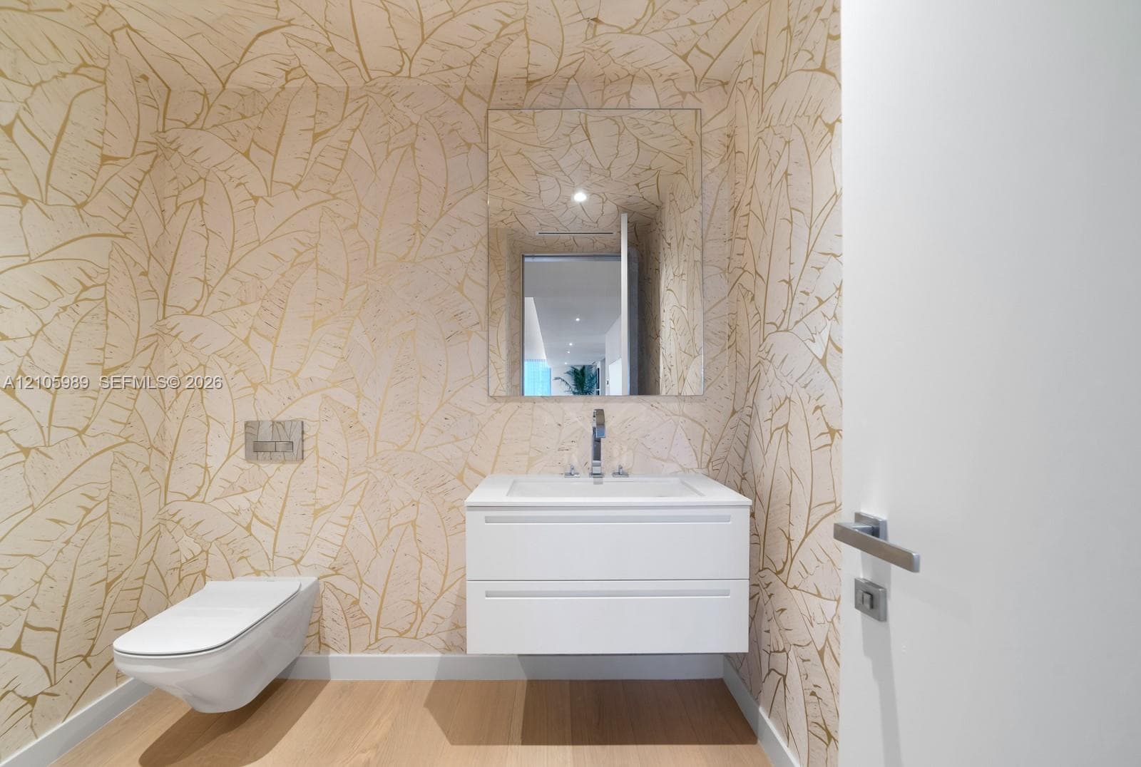 Floating white vanity with a rectangular sink, wall-mounted dual-flush toilet, large frameless mirror, wood-finish flooring, and metallic patterned wall covering
