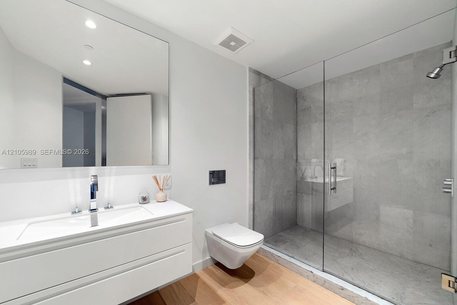 Floating white vanity with a rectangular sink, wall-mounted toilet, walk-in shower with a glass enclosure and stone-tile surround, and wood-finish flooring