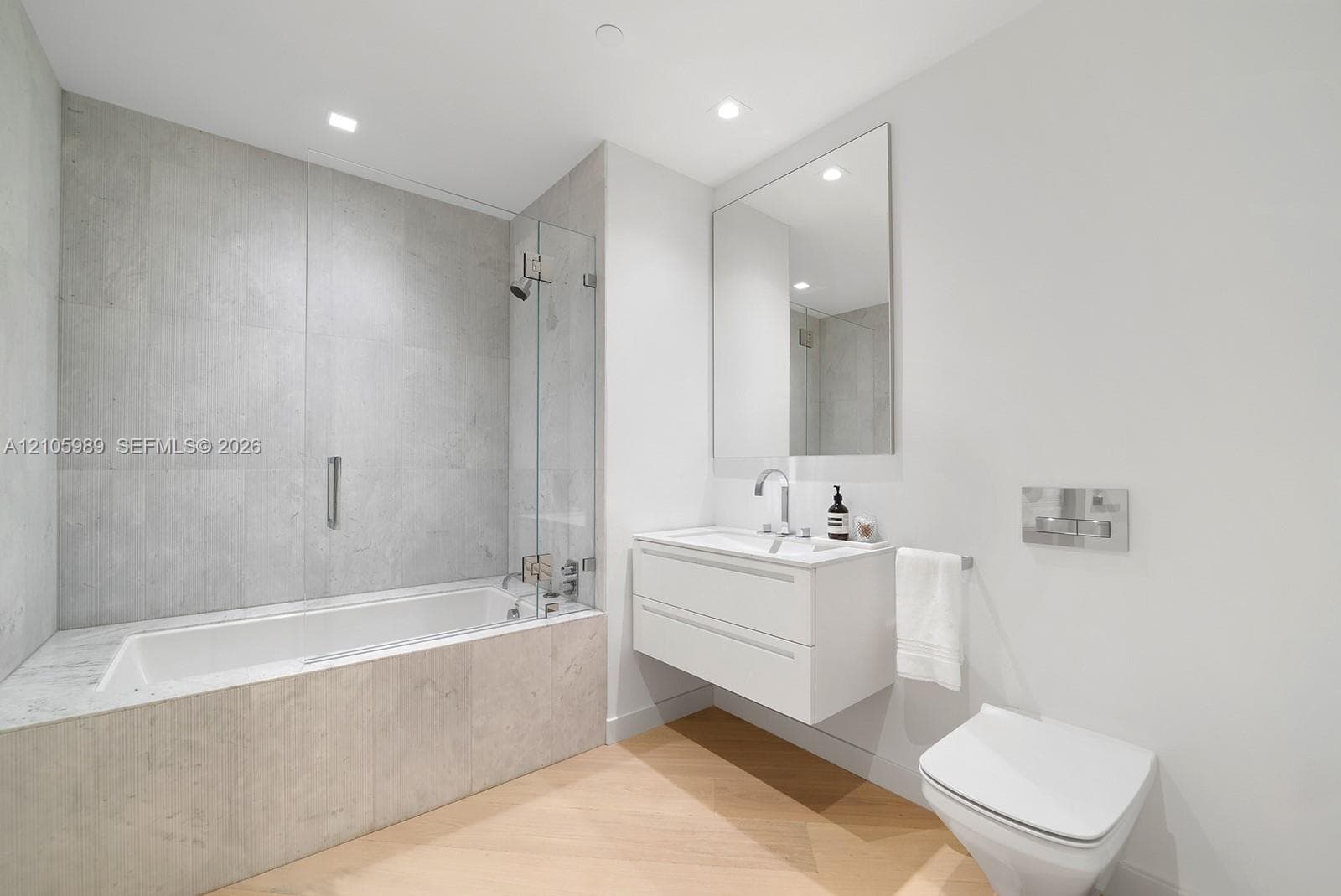 Bathroom featuring a white floating vanity, wall-mounted toilet with a chrome flush plate, glass-enclosed shower over tub combination, large frameless mirror, and light-toned wood-finish flooring