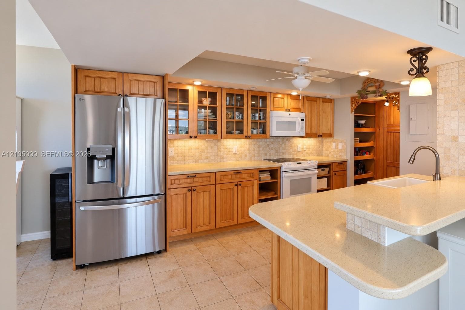 Kitchen featuring honey-toned wood cabinetry with glass-front uppers, solid surface countertops, a tile backsplash, stainless steel refrigerator, and a raised breakfast bar