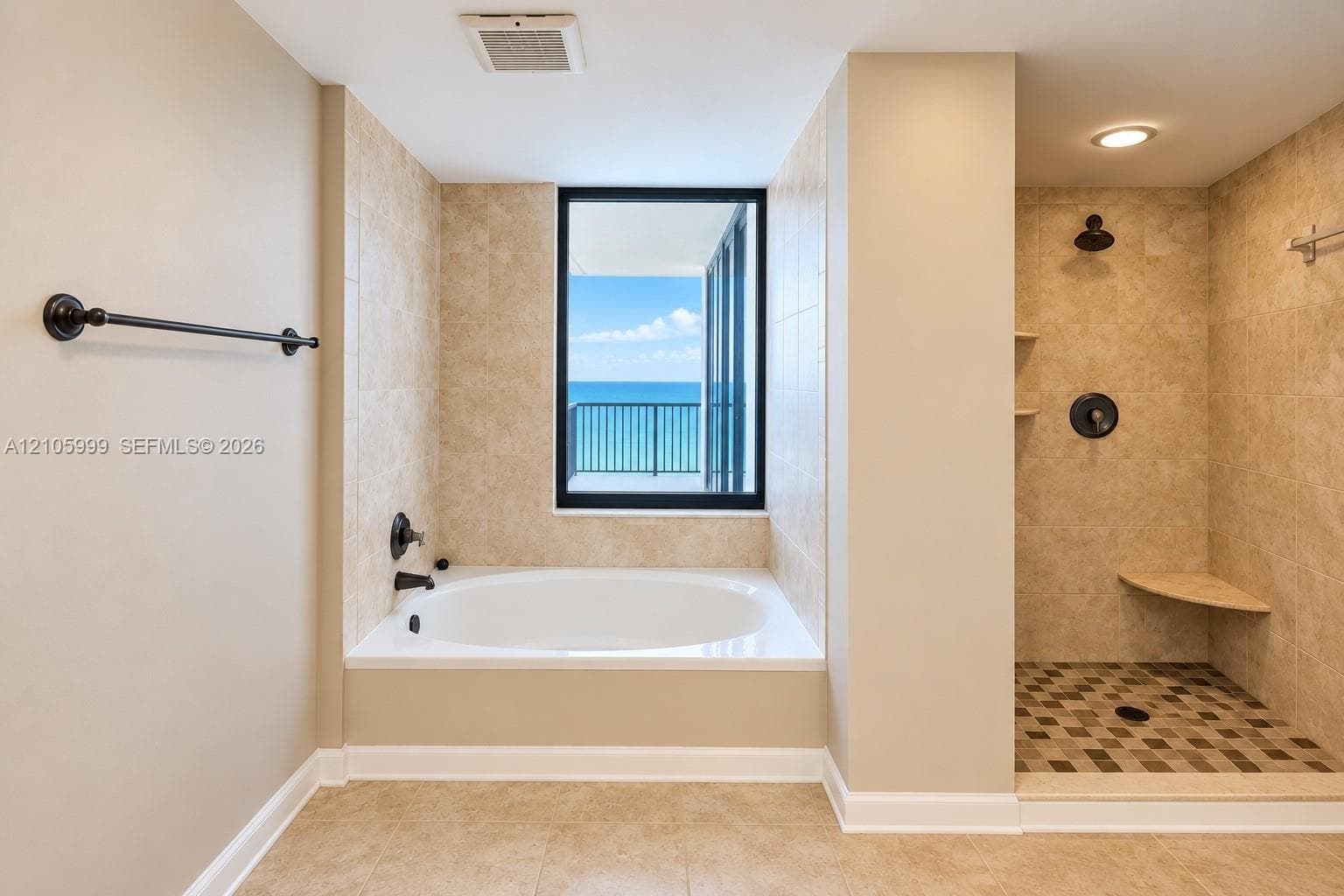 Deep soaking tub, walk-in shower with built-in bench seating, neutral stone-finish tile surround, dark bronze hardware fixtures, and exterior ocean backdrop
