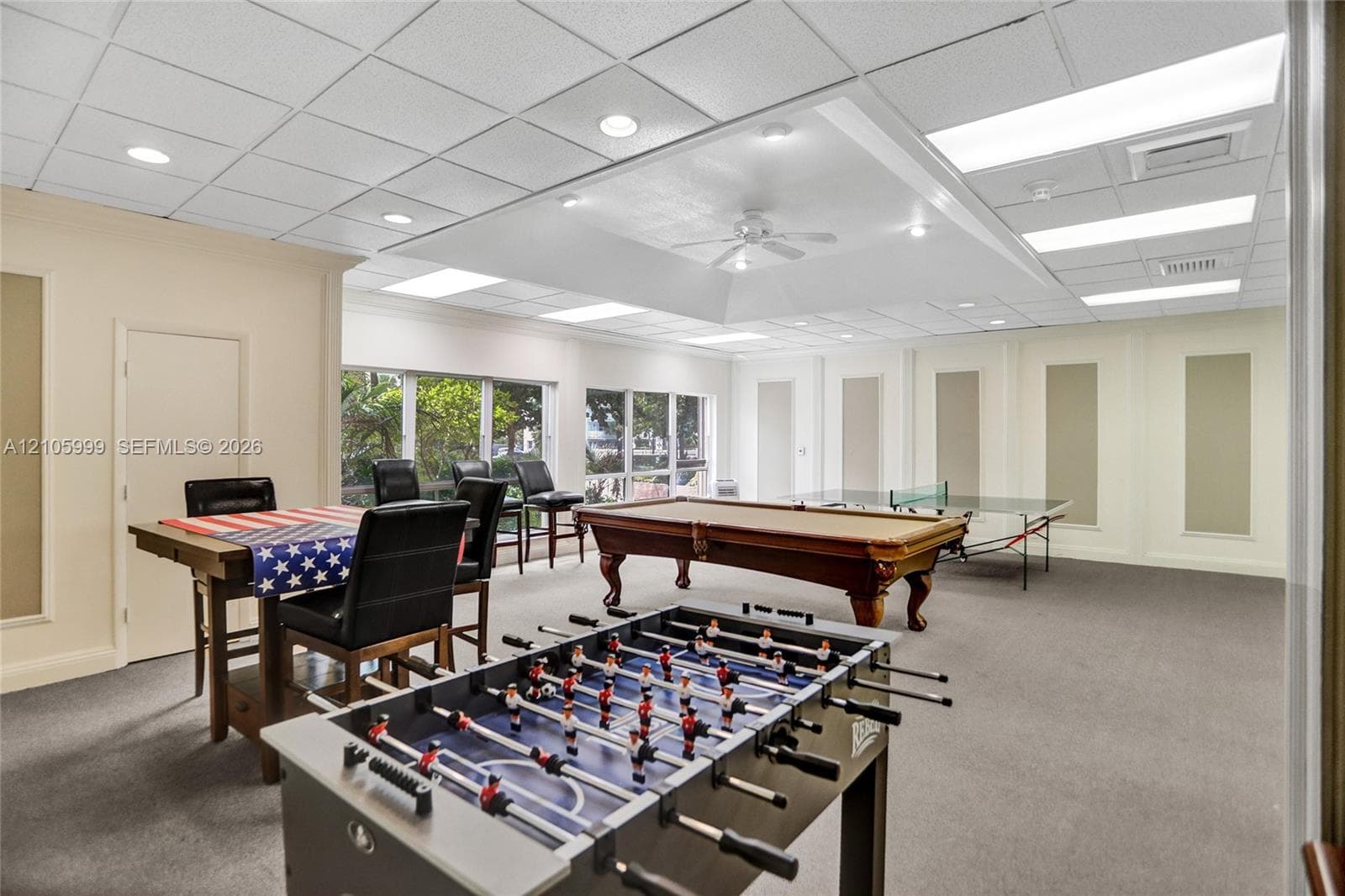 Large recreation space featuring a tiered ceiling with recessed lighting and a central ceiling fan, expansive wall-to-wall windows, and neutral carpeting