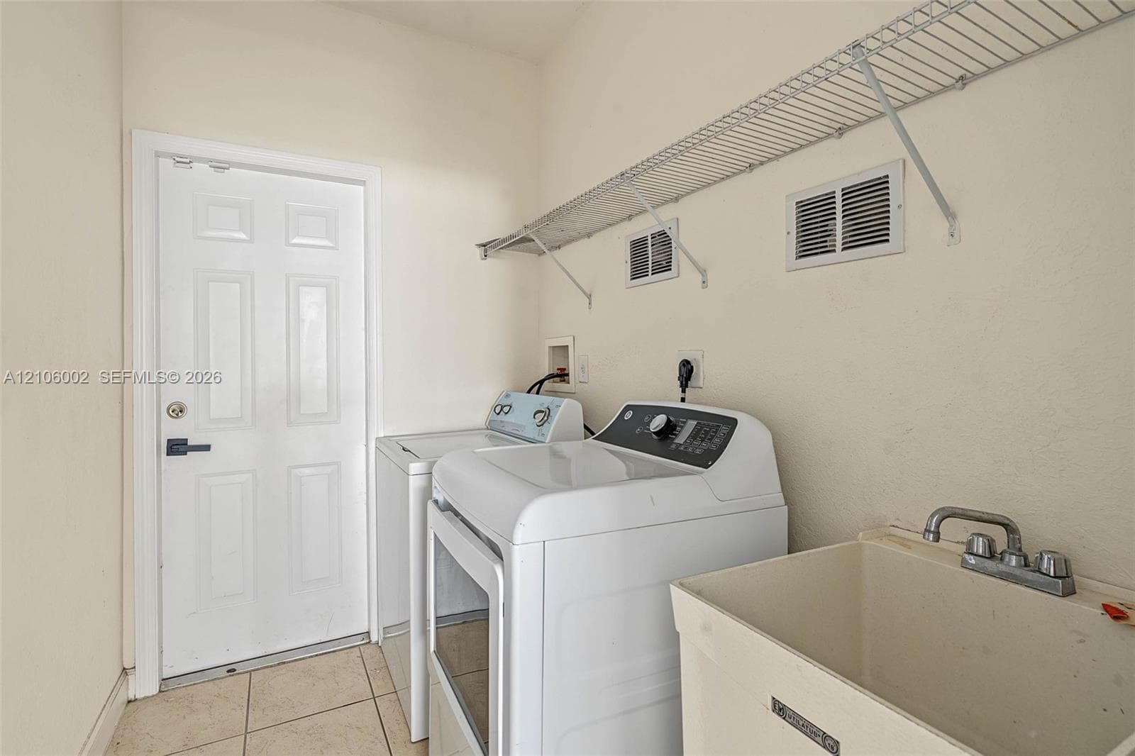 Utility room featuring tile flooring, a wall-mounted wire shelving unit, and a built-in utility sink with a dual-handle faucet