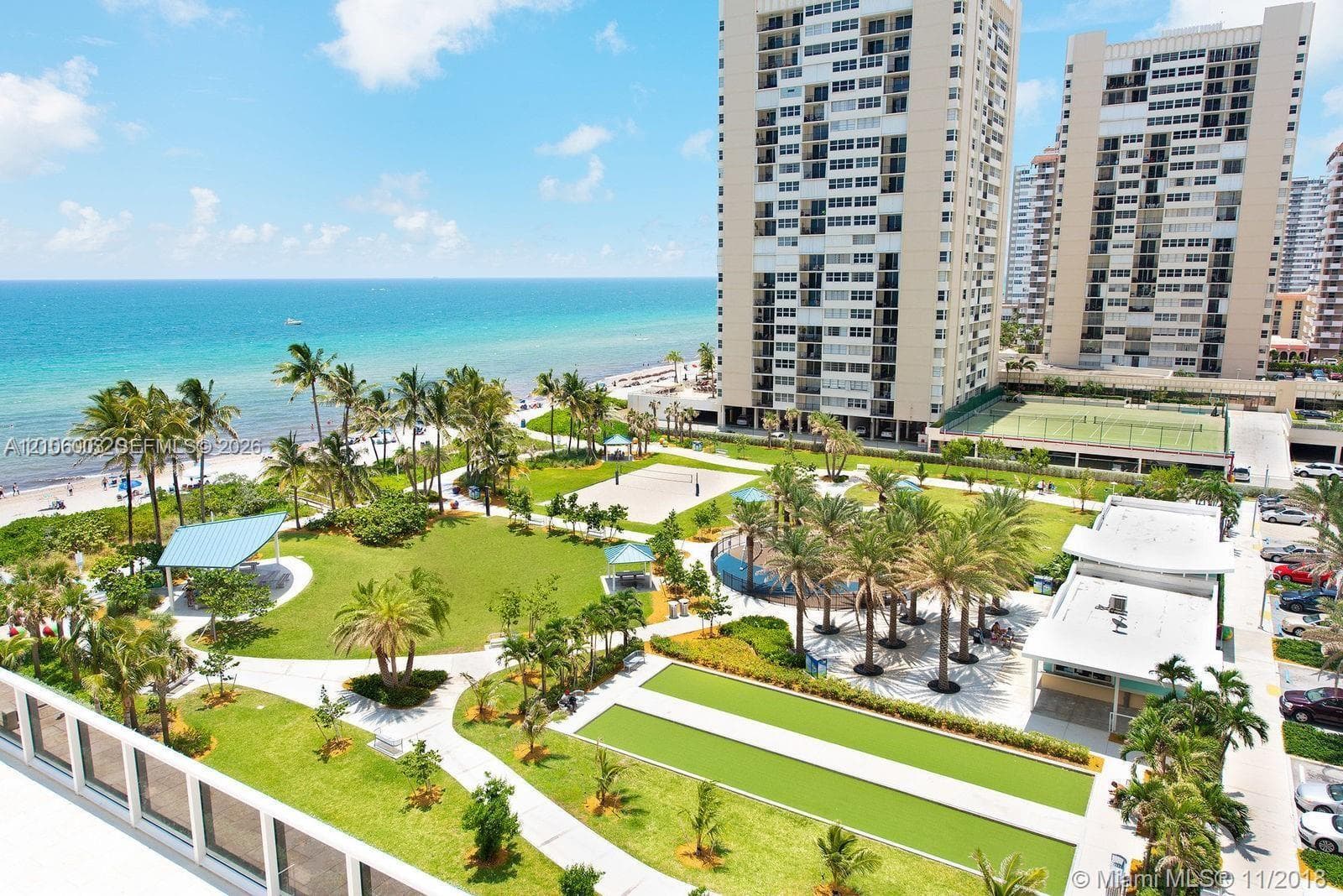 High-rise residential property featuring direct oceanfront access, landscaped green space, outdoor tennis courts, shaded pavilion areas, and palm-lined grounds