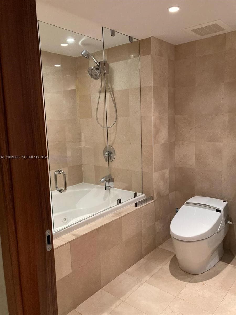 Bathroom featuring floor-to-ceiling stone-tile surround, soaking tub with a glass panel enclosure, hand-held shower fixture, and a one-piece low-profile toilet