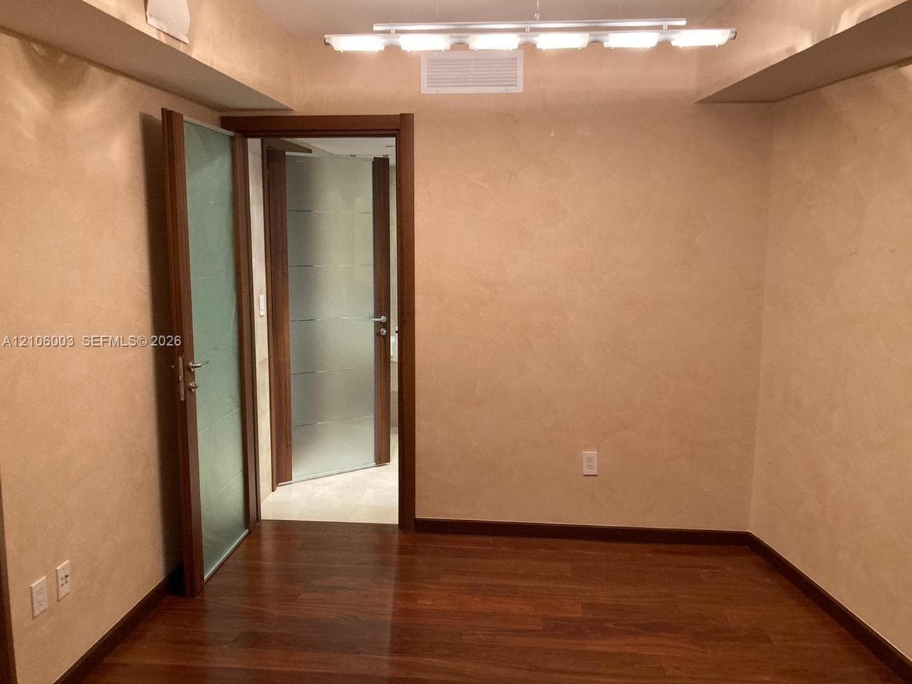 Wood-finish flooring with textured wall finishes, solid wood door frames, and frosted glass paneled doors featuring brushed metallic hardware, illuminated by a linear multi-light fixture