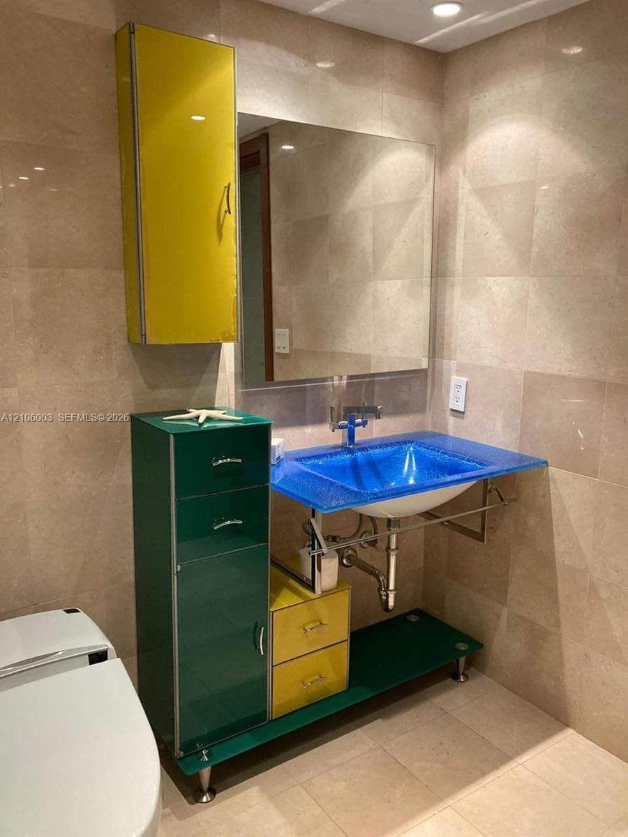 Bathroom featuring wall-to-wall light-toned stone tiling, a custom vanity with multi-colored high-gloss cabinetry, a blue glass basin sink, and a wall-mounted mirror