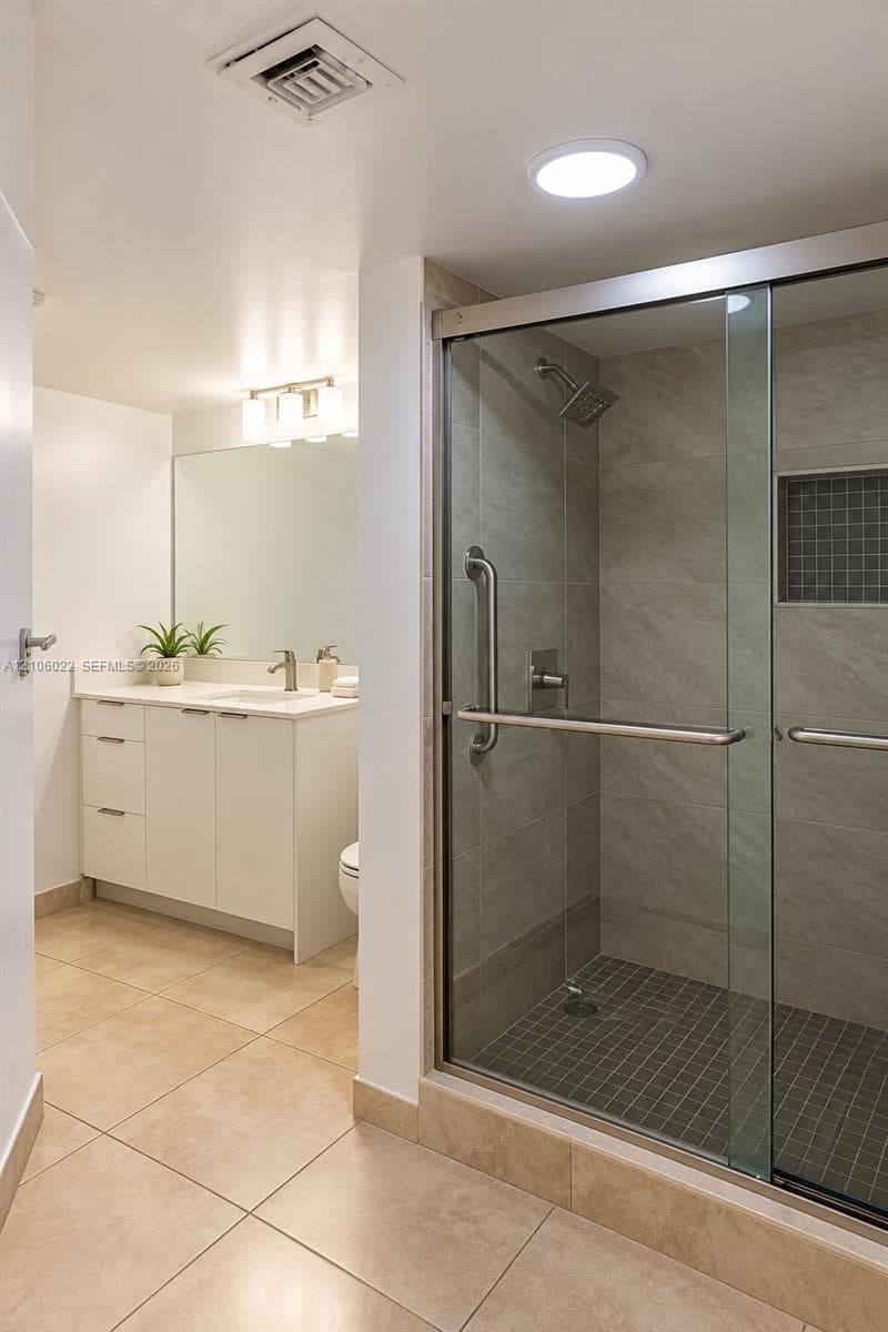 Bathroom featuring a white cabinetry vanity with integrated sink, shower enclosure with glass sliding doors, stone-finish tile surround, built-in shower niche, and large-format floor tiling