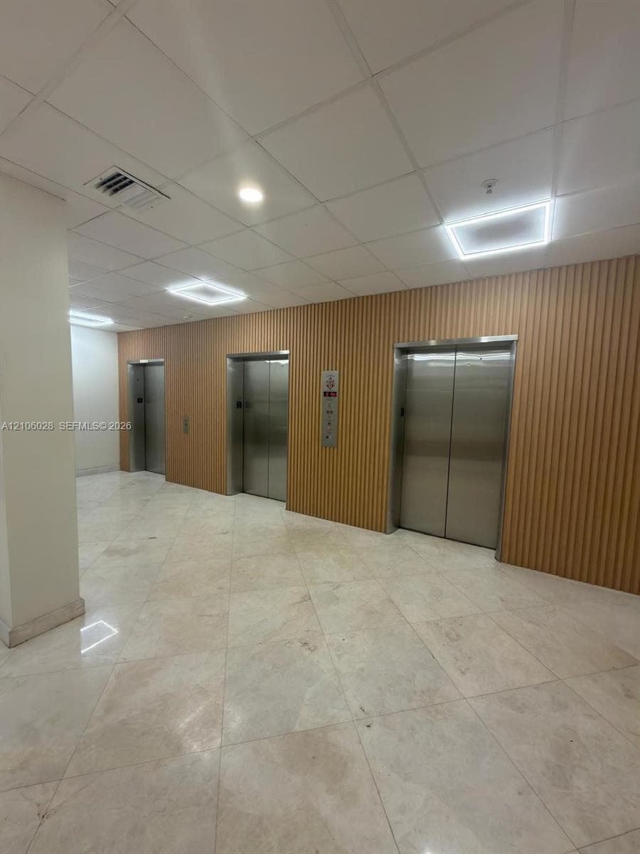 Elevator lobby featuring three stainless steel elevator doors, ribbed wood-panel wall cladding, polished stone flooring, and recessed LED ceiling panels