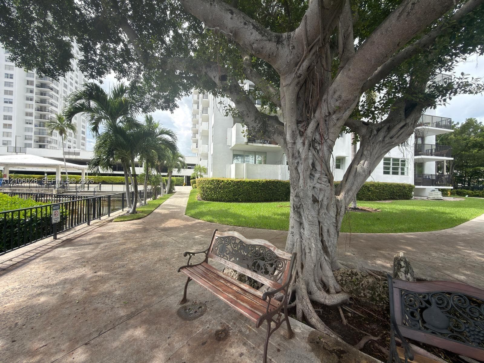Landscaped grounds featuring mature trees, manicured hedges, paved walking paths, and metal perimeter fencing