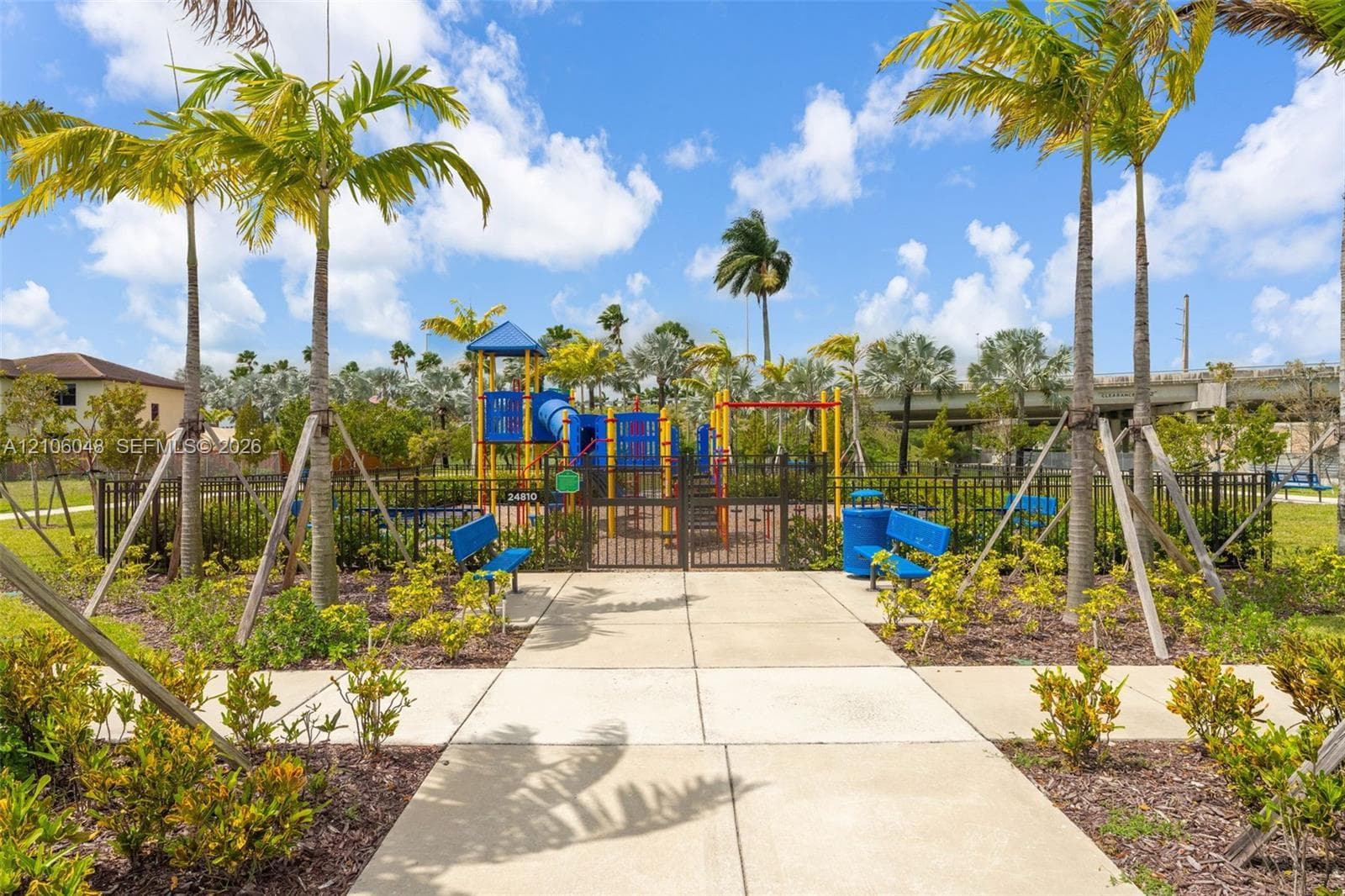 Fenced recreation area featuring a playground structure, concrete walking paths, built-in benches, and mature palm trees