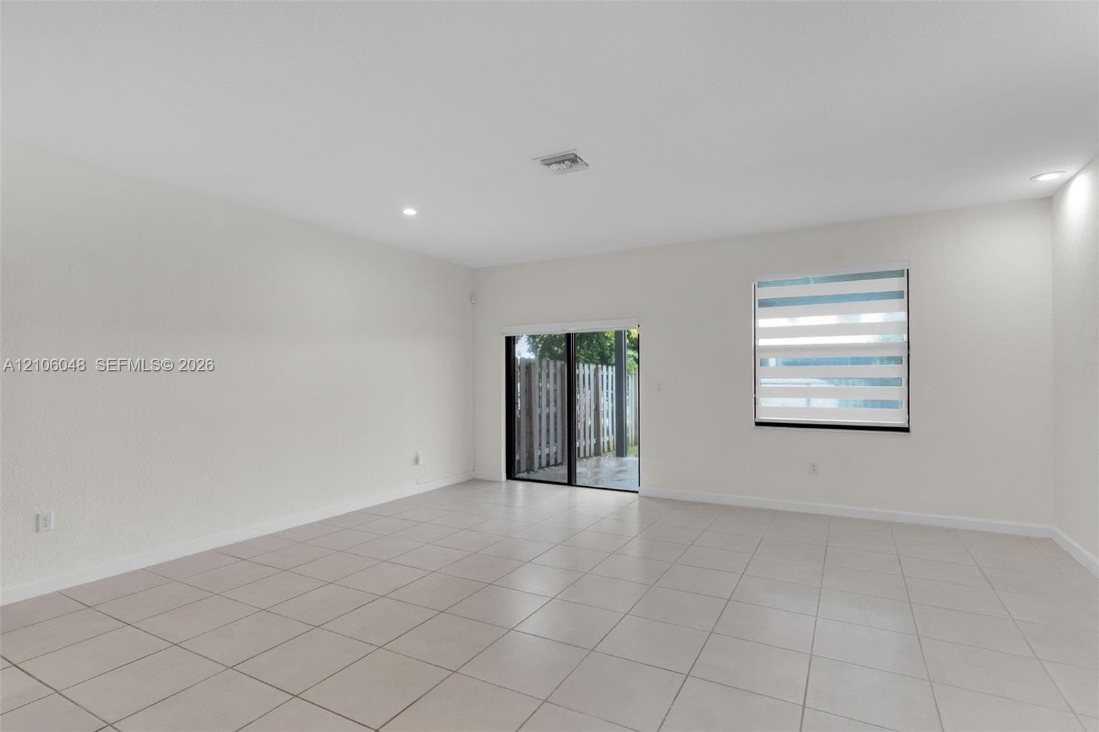 Neutral-toned interior space featuring light-colored ceramic tile flooring, recessed ceiling lighting, a single window with horizontal zebra-style blinds, and a sliding glass door