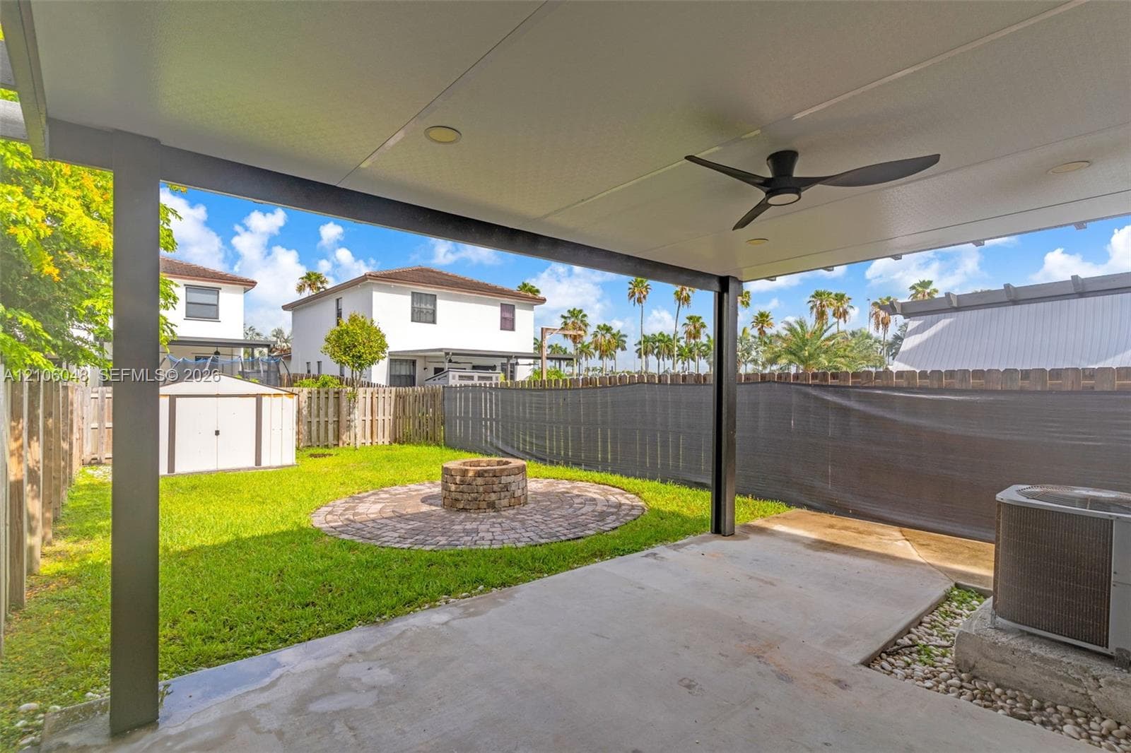 Covered concrete patio featuring a ceiling fan and recessed lighting, overlooking a fenced backyard with a circular stone fire pit