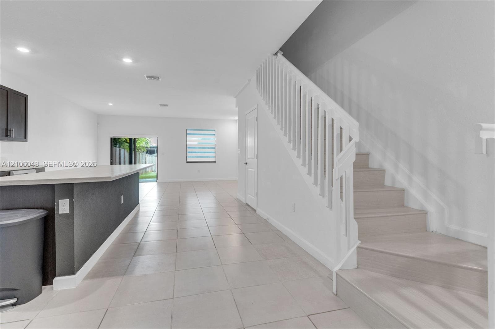 Interior featuring light-toned tile flooring, a kitchen island with stone-surface countertop, recessed ceiling lighting, and a staircase with white balusters