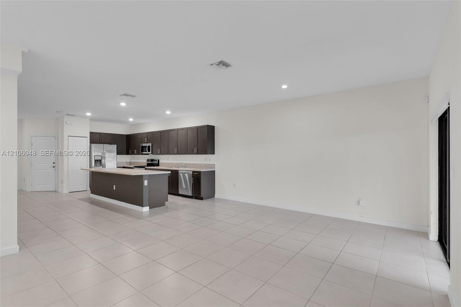 Open-concept kitchen featuring dark espresso cabinetry, a central island with countertop seating, stone-finish countertops, stainless steel appliances, and large-format tile flooring