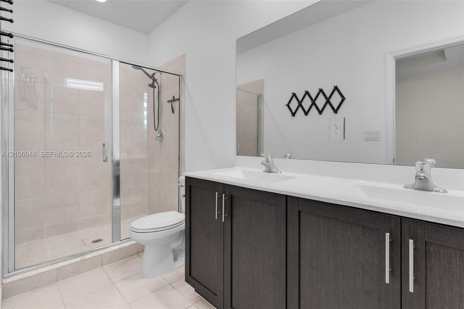 Double vanity with dark cabinetry, white countertops, integrated sinks, chrome hardware, and a glass-enclosed shower with tiled surround