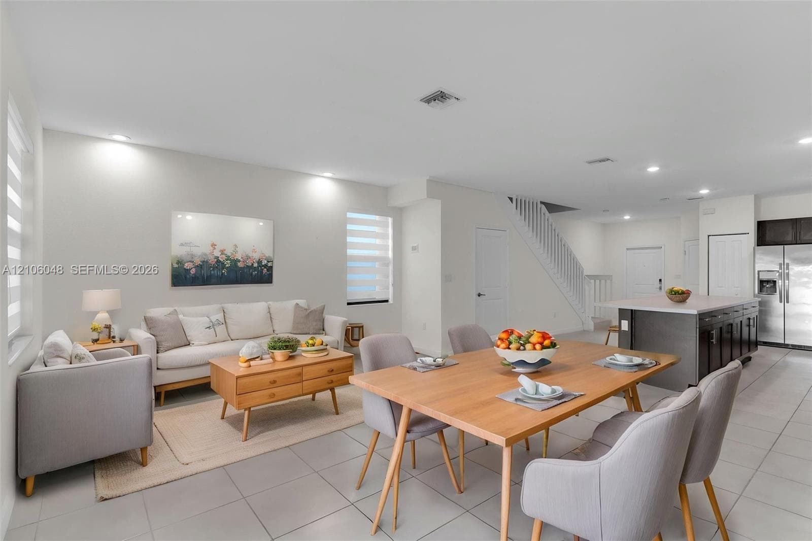 Open-concept interior featuring large-format tile flooring, recessed ceiling lighting, a kitchen island with stone countertops, and a white wooden staircase railing