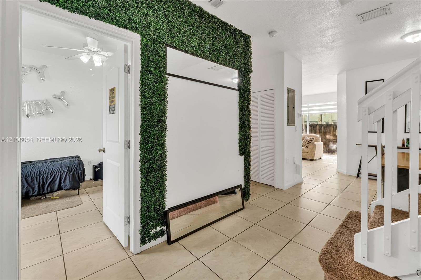 Interior space featuring large-format tile flooring, decorative faux-greenery wall accent, and white staircase with wooden balustrade