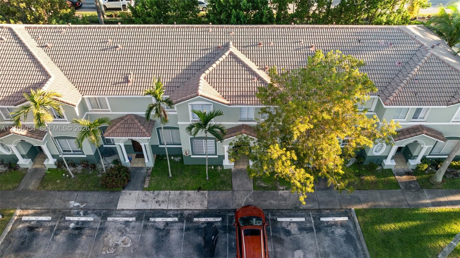 Multi-unit building exterior featuring a tile roof, light green stucco facade, decorative columns, arched entryways, and manicured palm trees