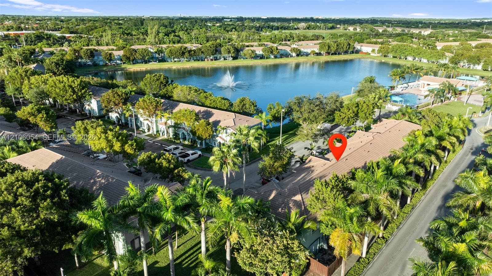 Aerial perspective featuring a multi-unit residential structure with tile roofing, situated within a landscape of mature palm trees and green space, positioned near a central pond with a water feature