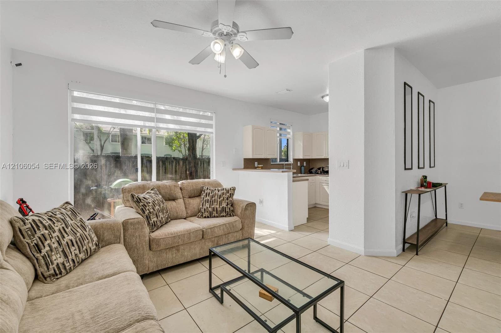Living area featuring large sliding glass doors, neutral tile flooring, ceiling fan with light kit, and pass-through counter to kitchen