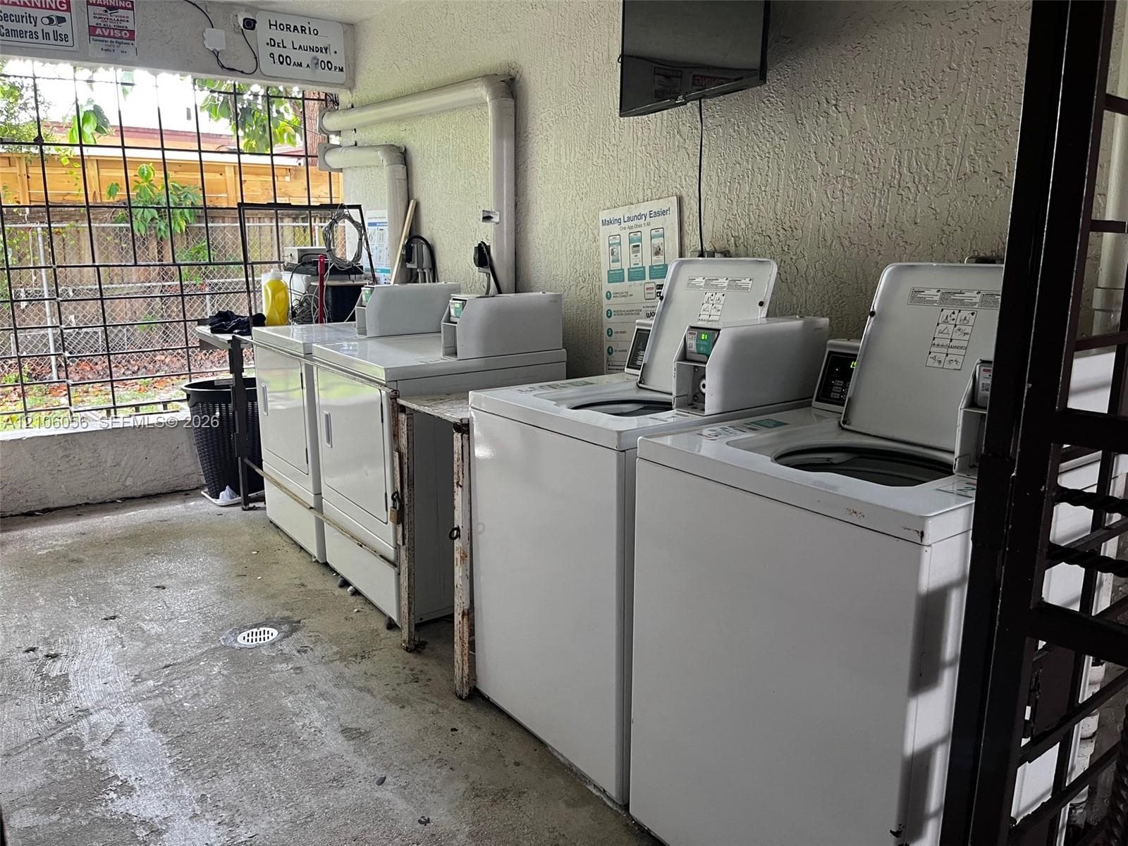 Laundry facility featuring multiple coin-operated washing machines, concrete flooring, exterior-facing metal security bars, and textured wall finish