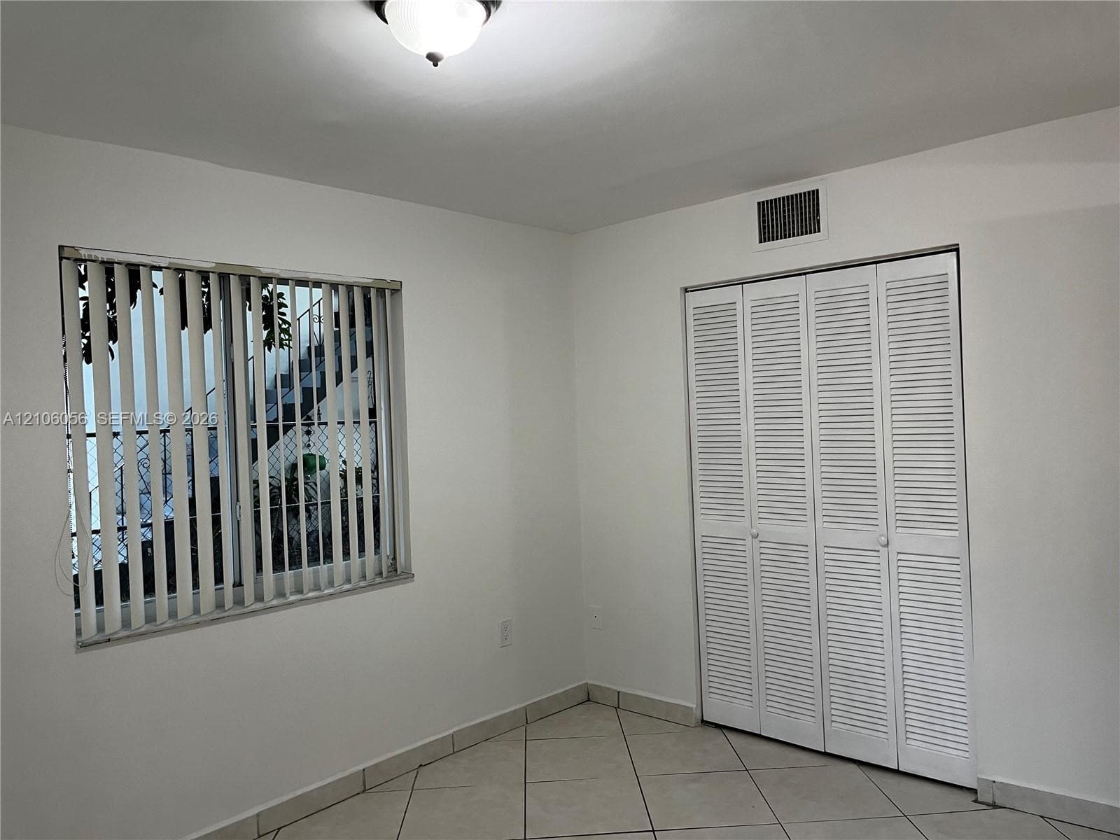 Interior space featuring neutral wall tones, large-format tile flooring, a window with vertical blinds, and a built-in closet with louvered bifold doors