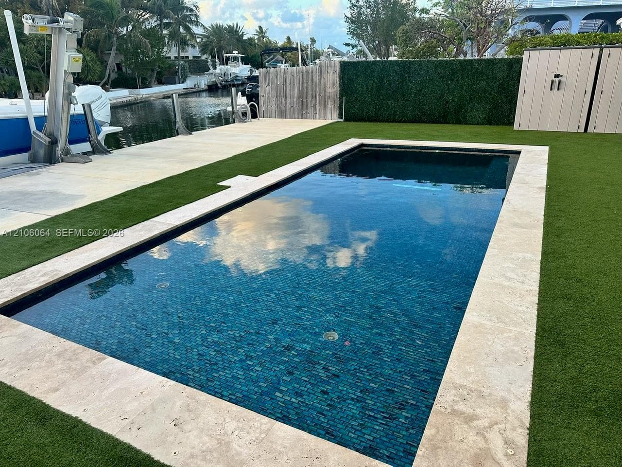 Rectangular pool with tile interior and natural stone coping, synthetic lawn surface, boat lift, concrete bulkhead, and wood-panel privacy fencing