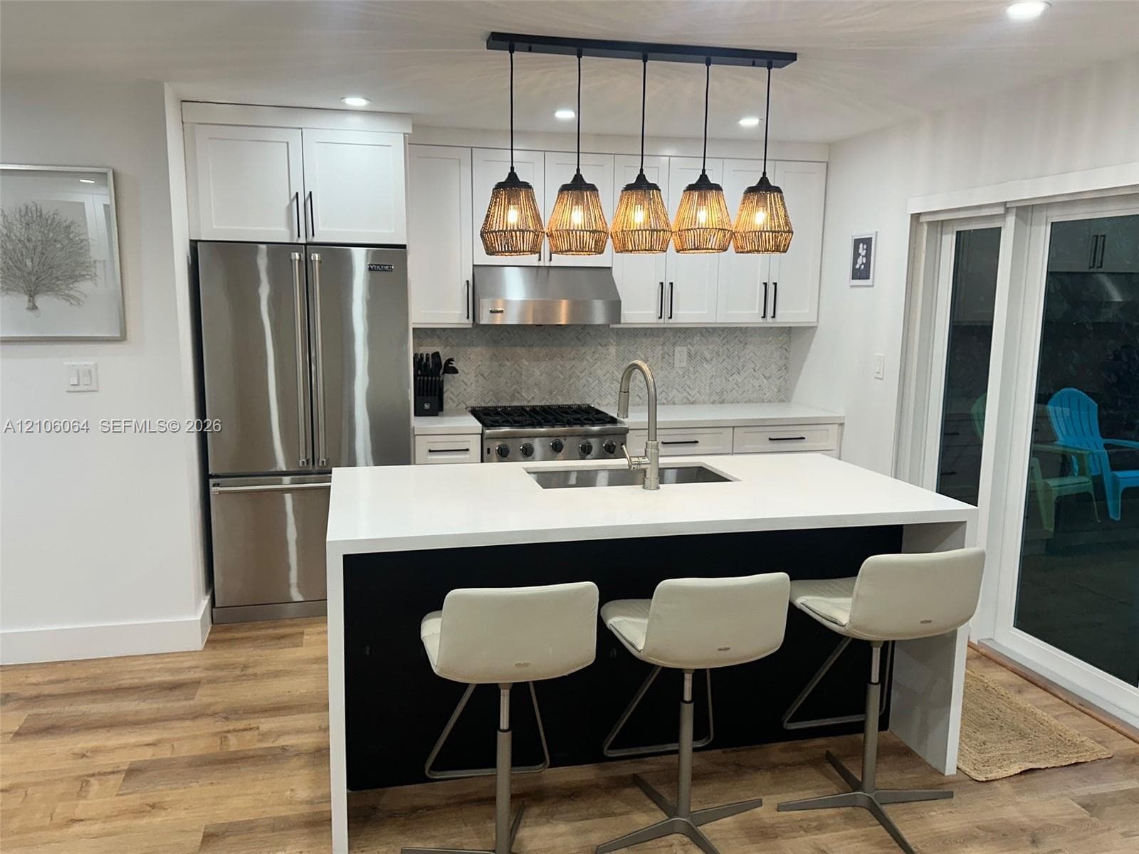 Kitchen featuring a central island with a solid surface countertop, white shaker-style cabinetry, stainless steel appliances, a herringbone tile backsplash, and a five-light hanging pendant fixture