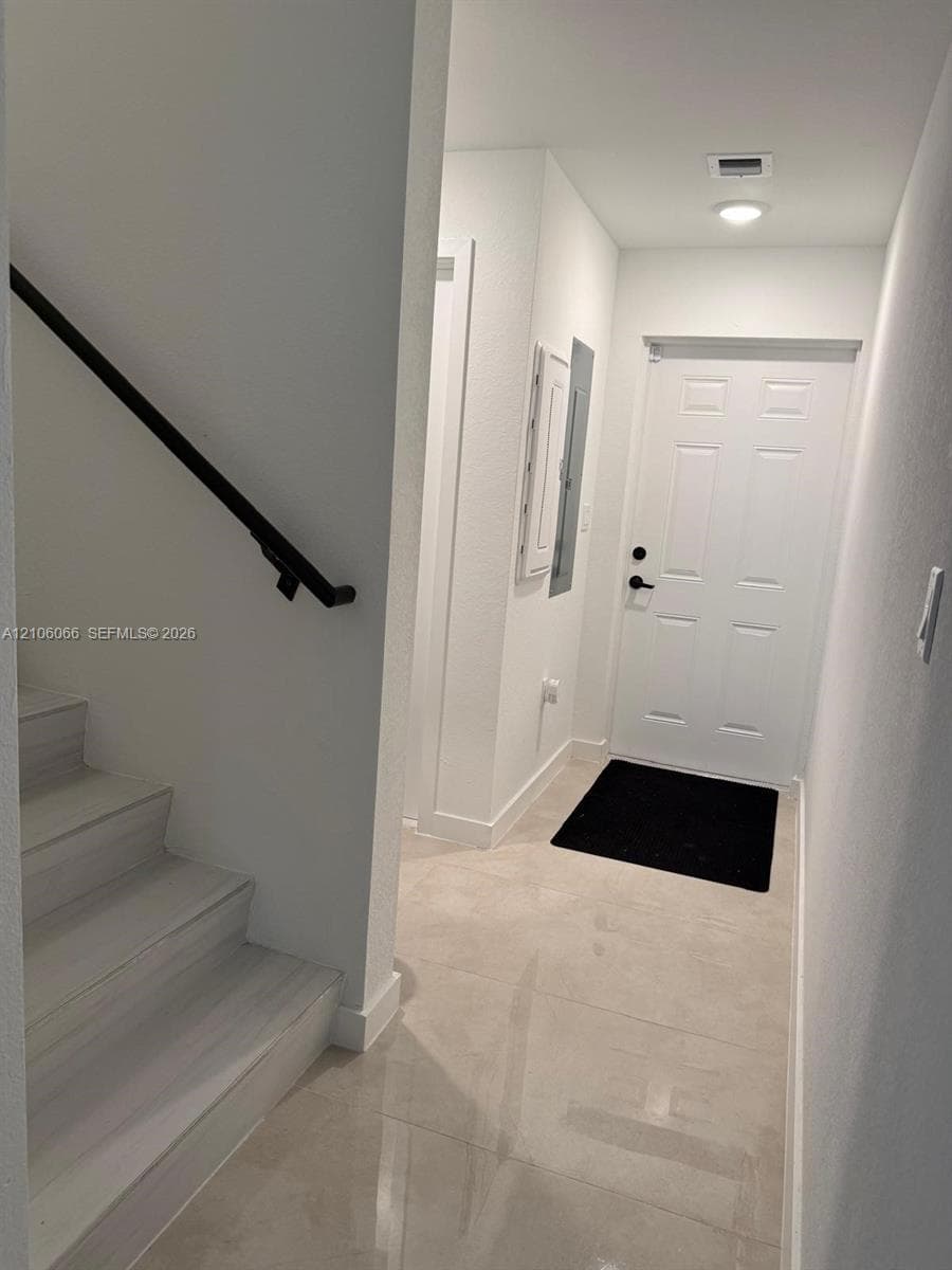 Entryway featuring large-format neutral tile flooring, a six-panel exterior door with matte black hardware, and a staircase with wood-finish treads and a matte black wall-mounted handrail