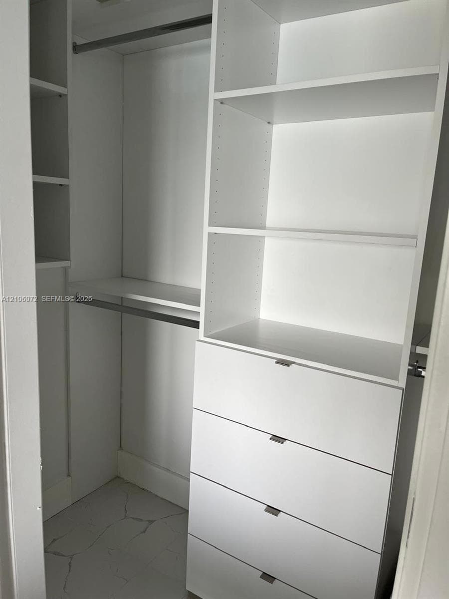 Custom closet featuring white built-in shelving, a four-drawer dresser unit, metal hanging rods, and stone-finish flooring
