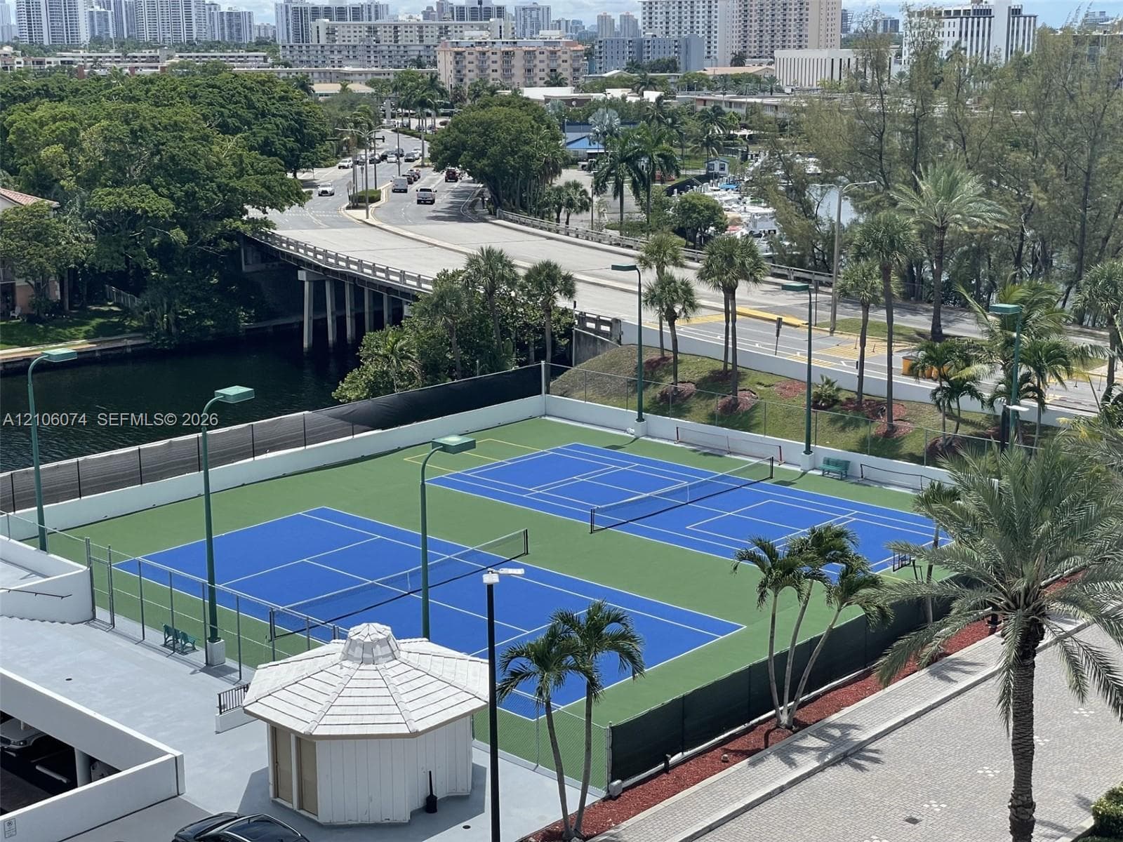 Dual tennis courts featuring hard-surface playing areas, perimeter fencing, overhead lighting, and a central gazebo