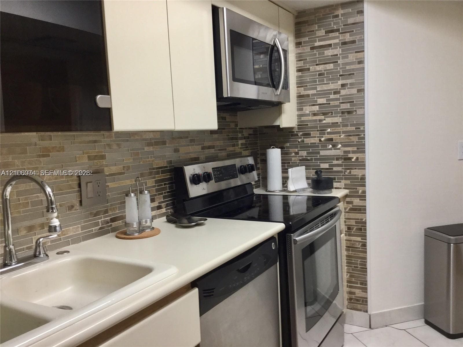Kitchen featuring stainless steel appliances, multi-tonal linear glass tile backsplash, solid-surface countertops, dual-basin sink with arched faucet, and flat-panel cabinetry