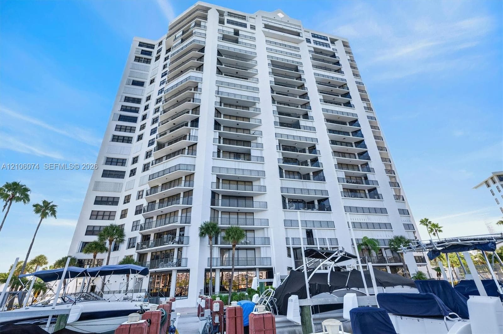High-rise building exterior featuring tiered private balconies, glass railings, and a concrete facade, set against a marina with boat docking slips