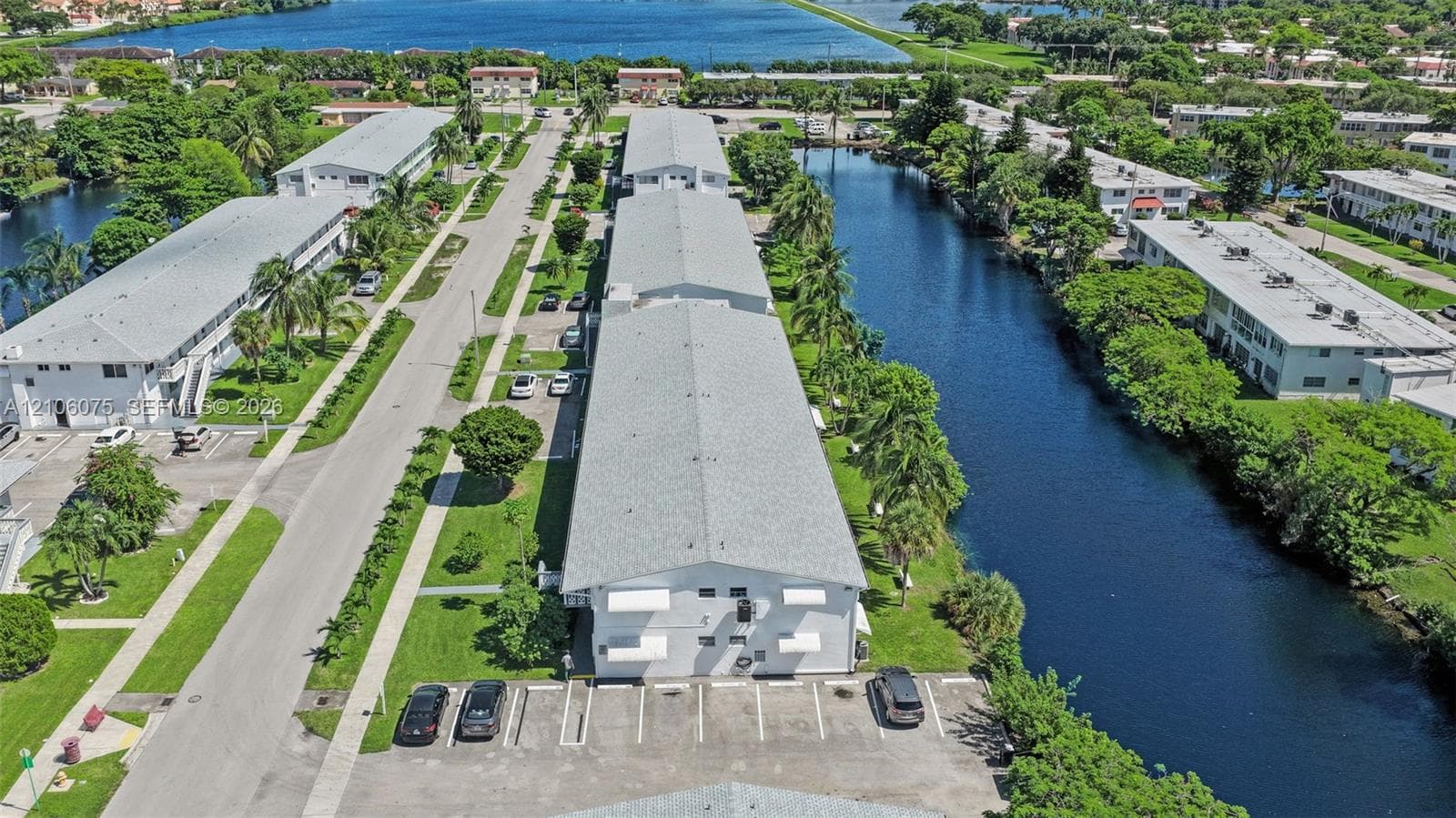 Aerial perspective of multi-family property featuring canal-front positioning, asphalt parking areas, shingled roofing, and surrounding mature landscaping