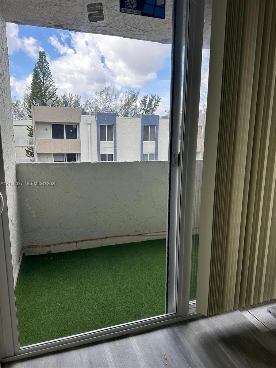 Private balcony featuring artificial turf flooring and a stucco perimeter wall, accessed through sliding glass doors with vertical blinds