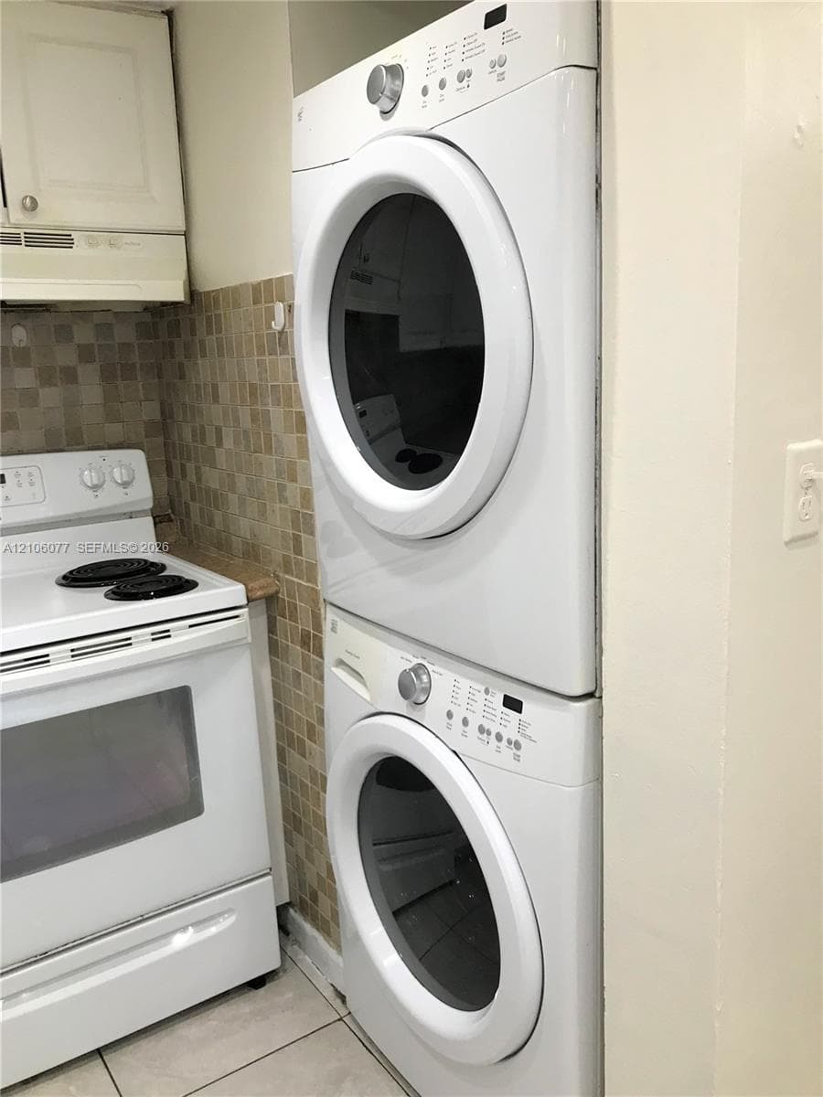 Stacked laundry unit, white electric range with four-burner cooktop, upper cabinetry, and mosaic tile backsplash