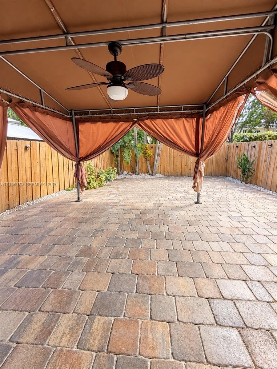 Outdoor patio featuring multi-tonal concrete pavers, a metal-framed overhead canopy, and a built-in ceiling fan, enclosed by a full-perimeter wood privacy fence