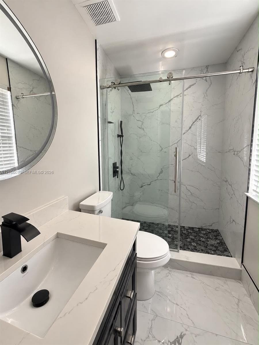 Dark-finish vanity with a light-toned countertop, integrated basin, matte black faucet, and frameless glass shower enclosure featuring large-format stone-patterned wall tile and a pebble-stone floor