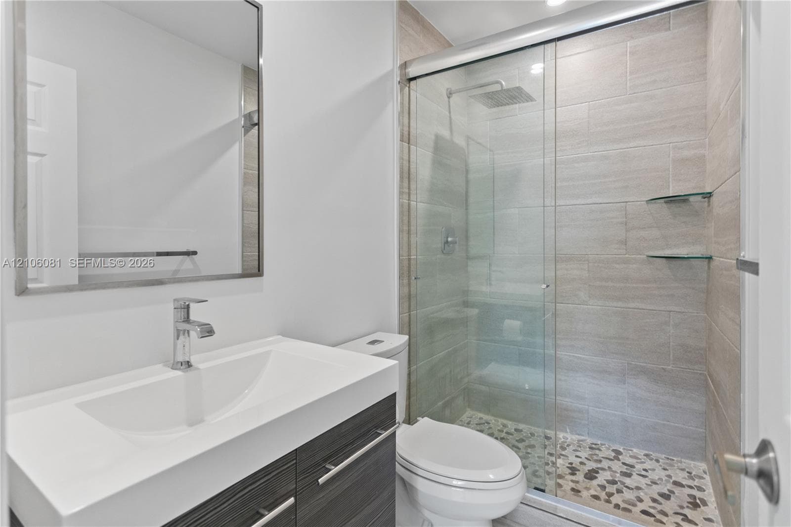 Bathroom featuring a floating vanity with a white countertop, wall-mounted mirror, glass-enclosed shower with tile surround, pebble-stone flooring, and a rainfall showerhead