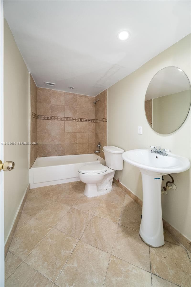 Full bathroom featuring neutral tile flooring, a pedestal sink, a circular wall-mounted mirror, and a bathtub with tile surround