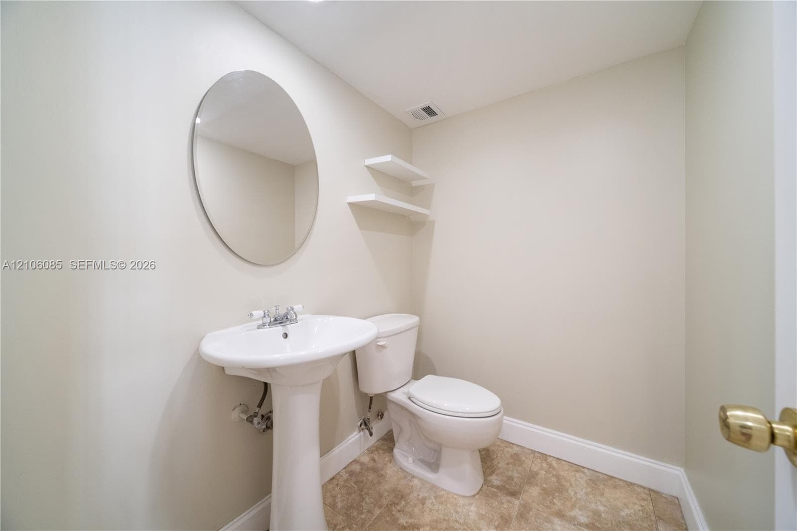 Ceramic pedestal sink with chrome faucet, oval wall-mounted mirror, two floating wall shelves, and tile flooring